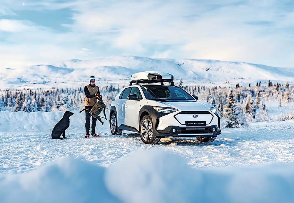 En vit Subaru Solterra med takbox i vinterlandskap med man och hund vid sidan av bilen.