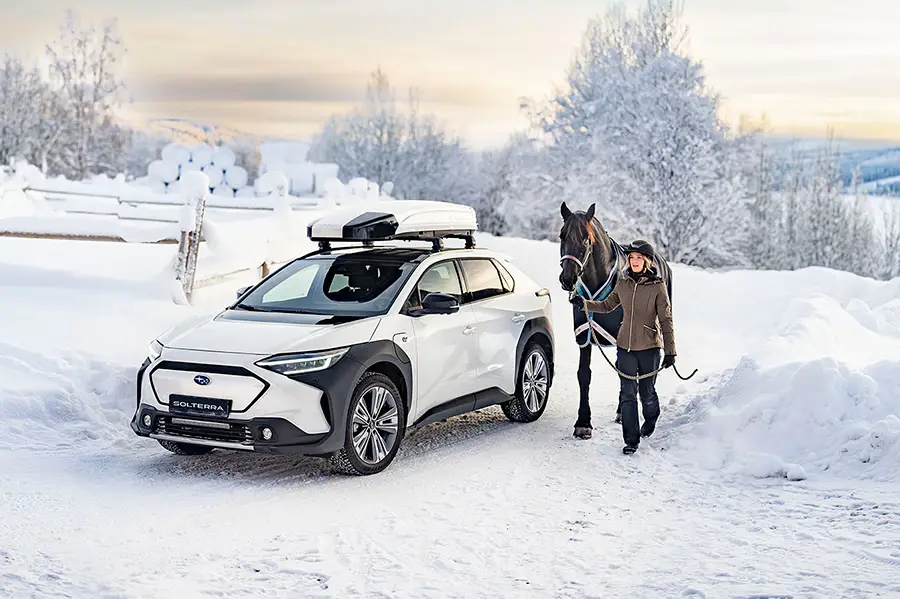 En vit Subaru Solterra med takbox i vinterlandskap med tjej som leder en häst brevid bilen.