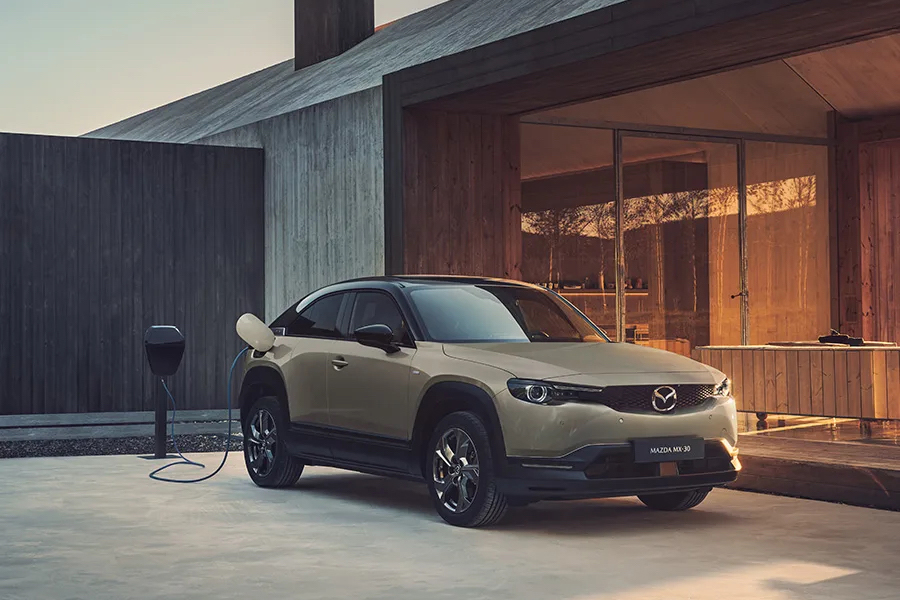 Beige Mazda MX-30 med inkopplad laddkabel parkerad vid ett arkitektritat hus