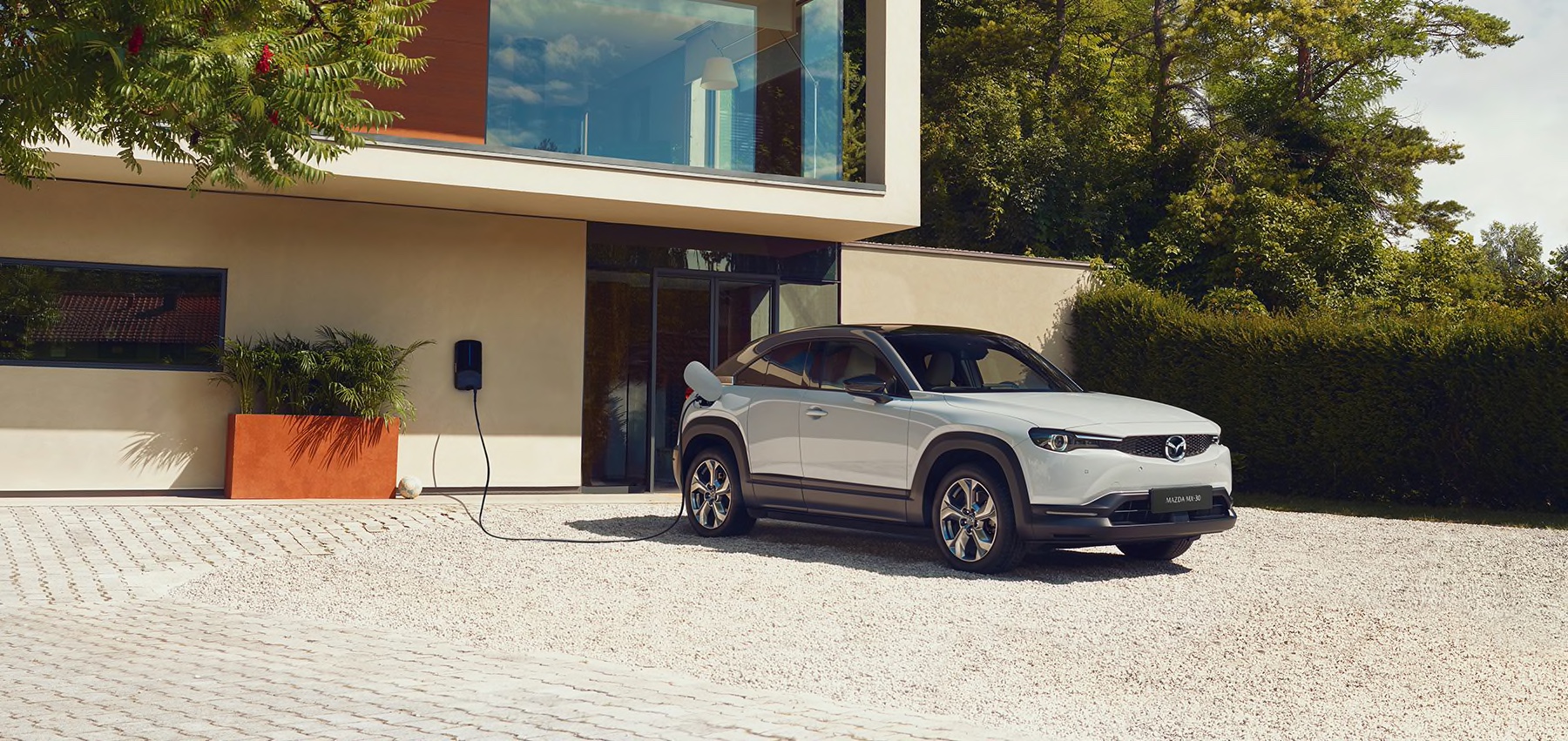 Vit Mazda MX-30 med inkopplad laddkabel parkerad vid ett arkitektritat hus