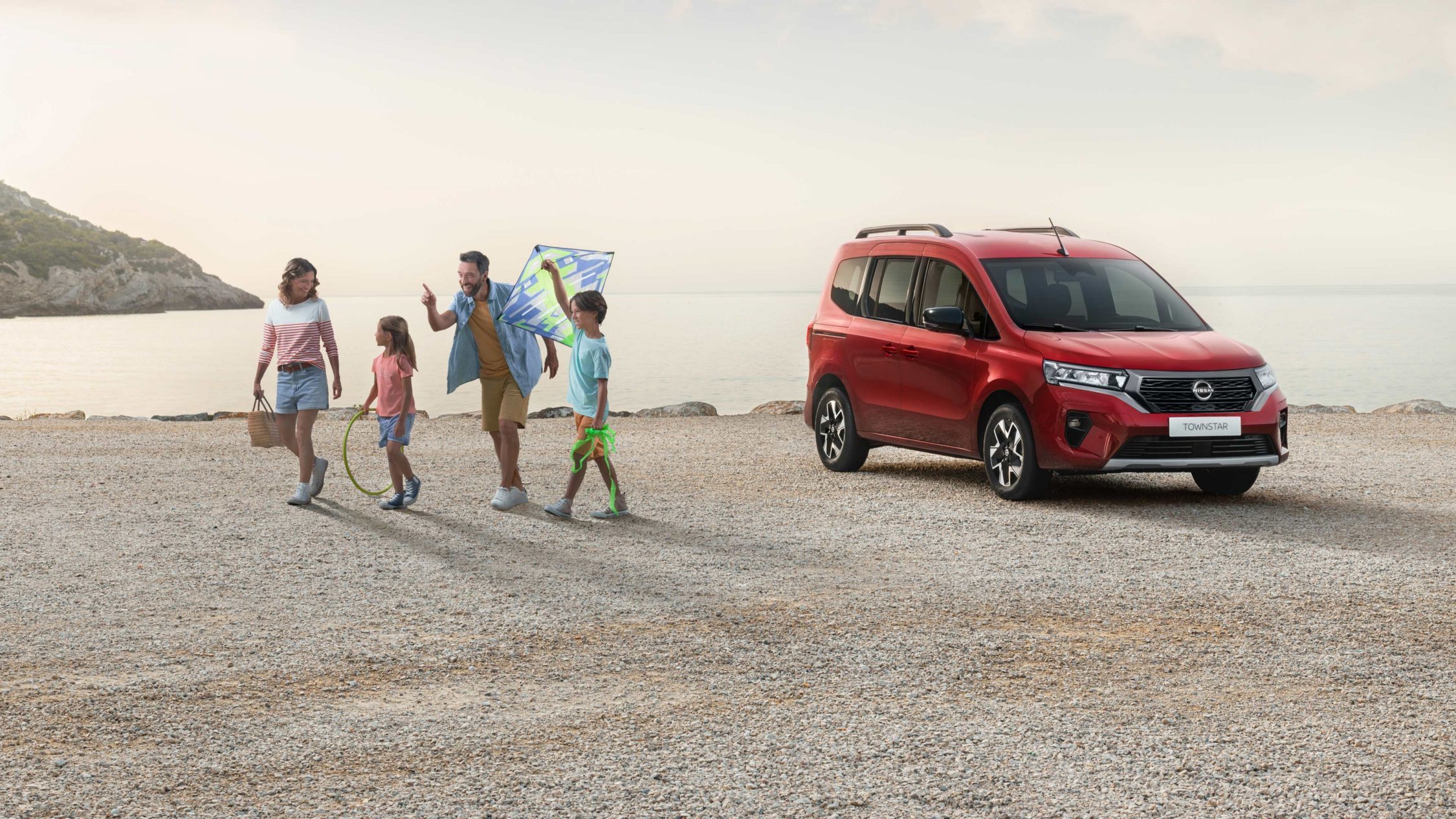Röd Nissan Townstar Combi parkerad vid havet och familj som flyger drake
